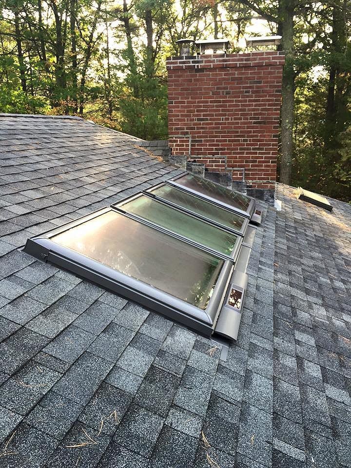 Skylight on a shingled roof next to a brick chimney in a wooded setting.