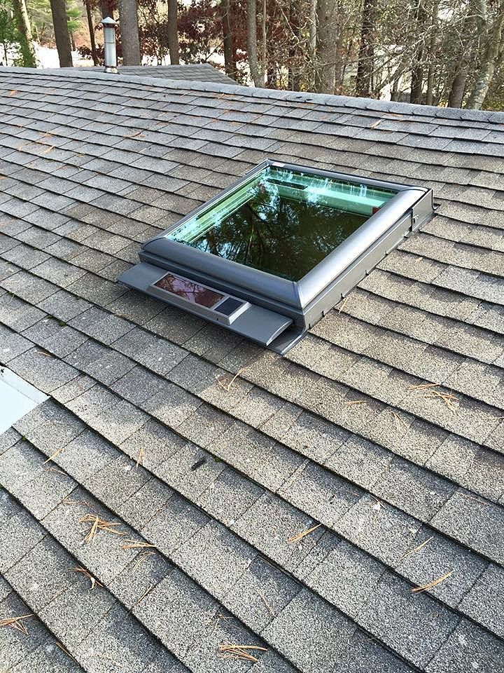 Skylight on a shingled roof, set against a backdrop of trees and a bright sky.
