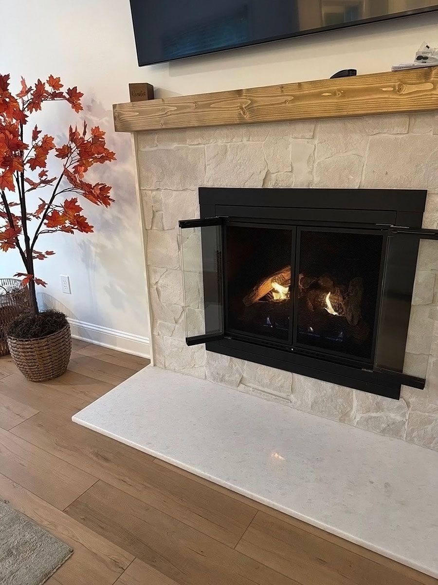 Living room fireplace with a black screen, lit fire, white hearth, wood mantel, and a potted plant nearby