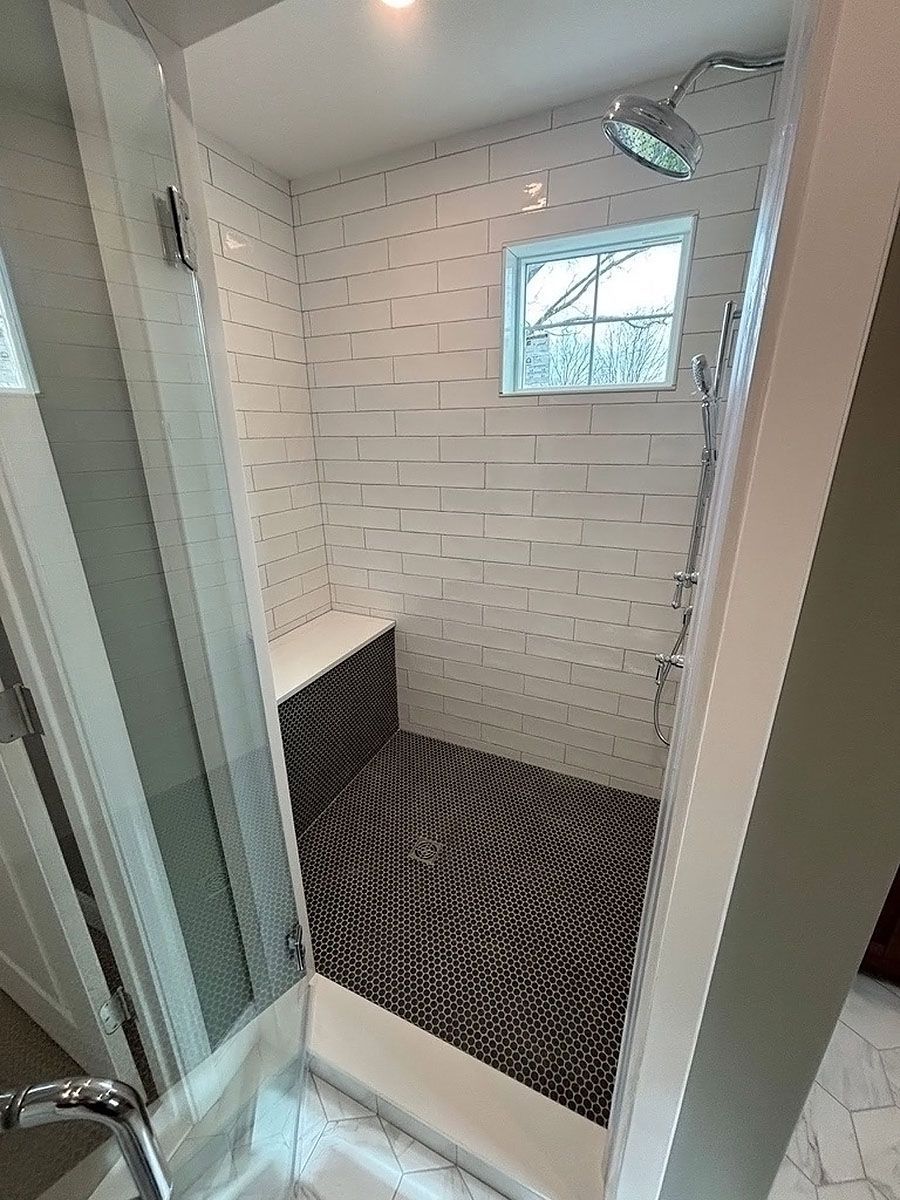 Small tiled shower stall with a glass door, white walls, and a black patterned shower mat.