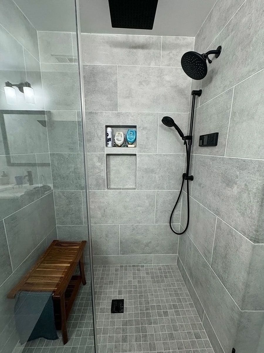Modern tiled shower stall with glass door, black showerhead, built-in niche, and wooden bench.