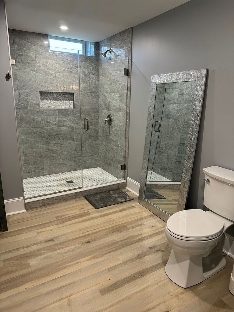 Modern bathroom with tiled shower, glass panel, large mirror, wood floor, and toilet
