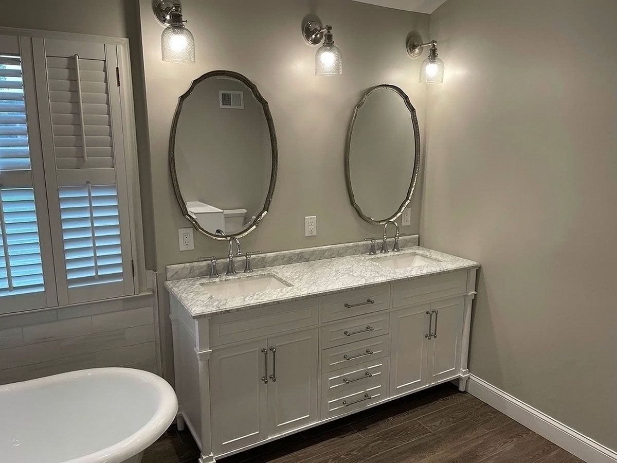 Bathroom vanity with two oval mirrors, wall sconces, and a white marble countertop beside a window shutter.