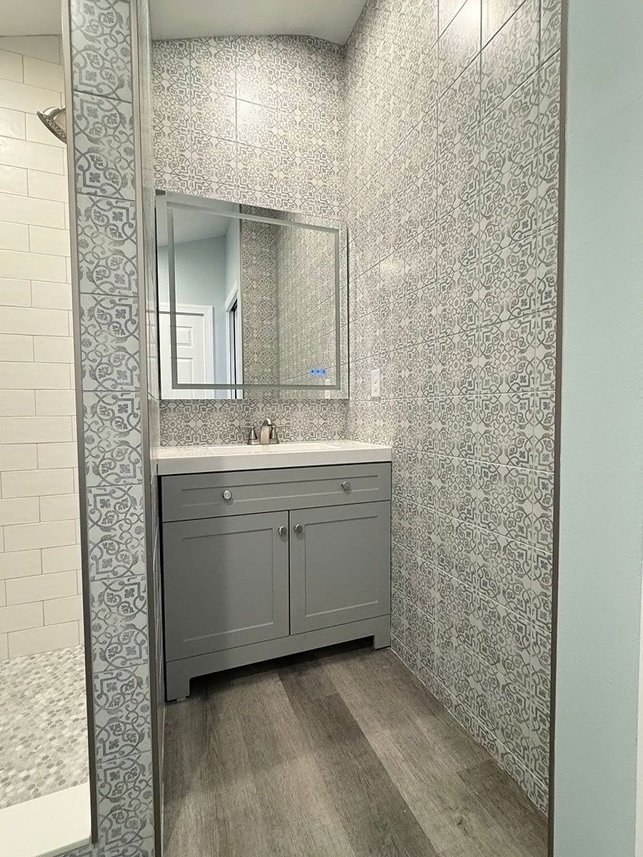 Small bathroom vanity with mirror and gray cabinet in a narrow wallpapered alcove.