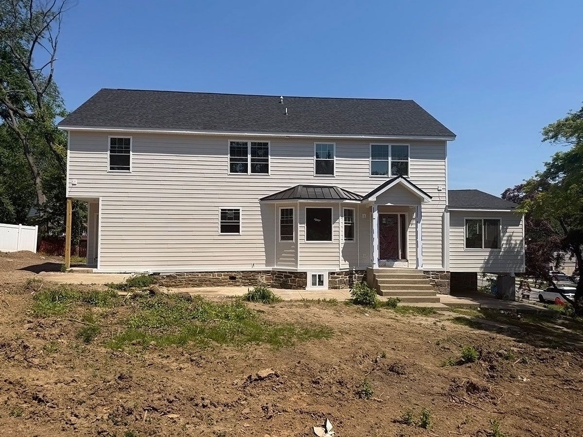 Two-story beige house with dark roof and front porch, on a bare yard under a clear blue sky