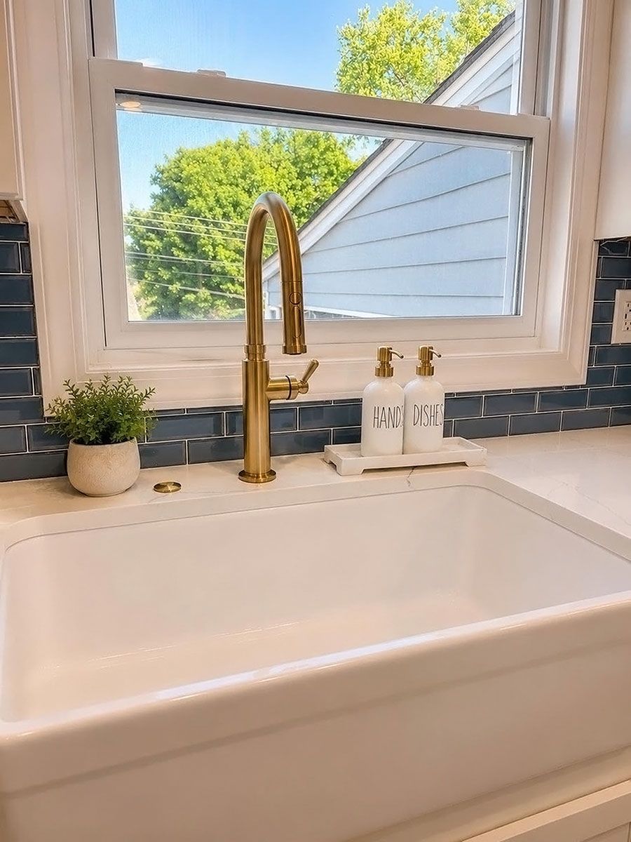 White farmhouse sink with gold faucet by a window, small plant and soap bottles on the ledge.