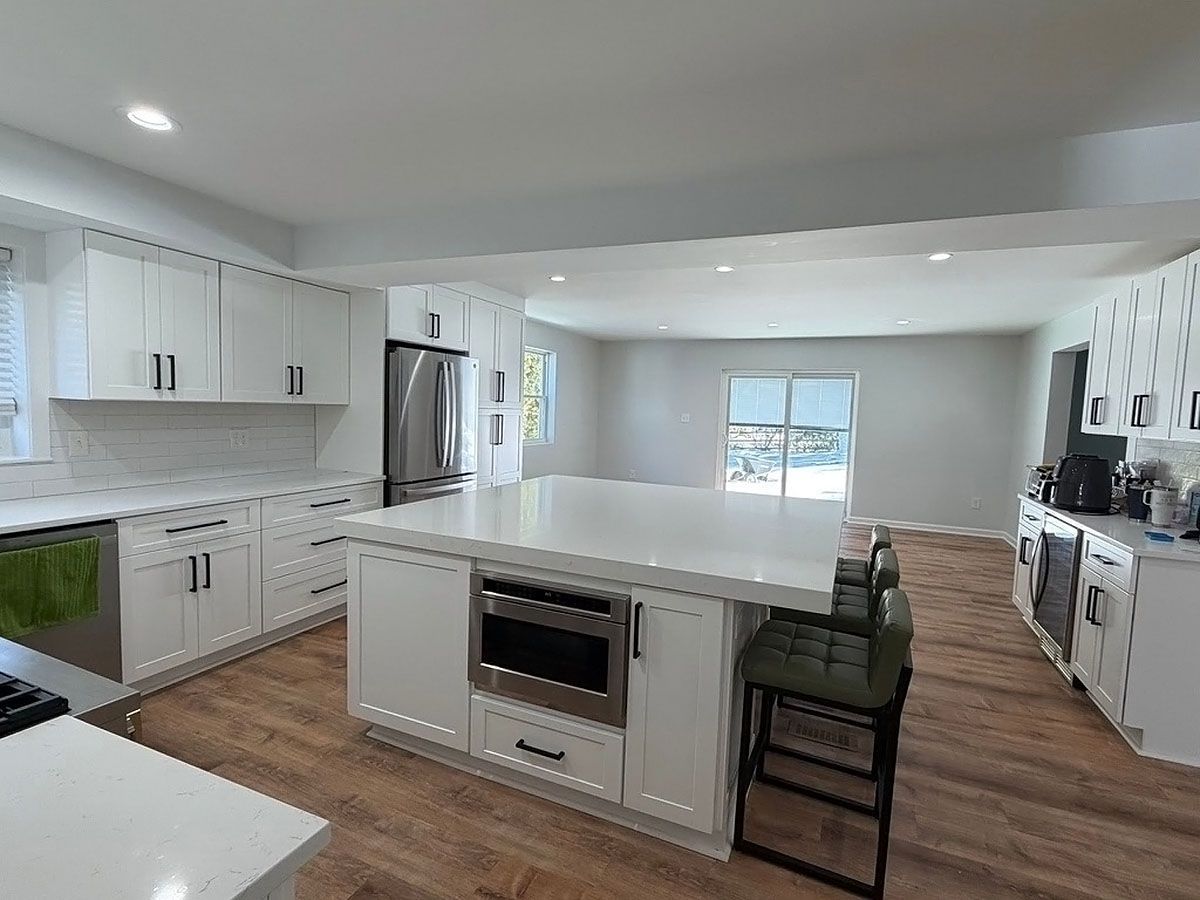 Bright white kitchen with large island, stainless appliances, wood floors, and a view to a windowed living area.