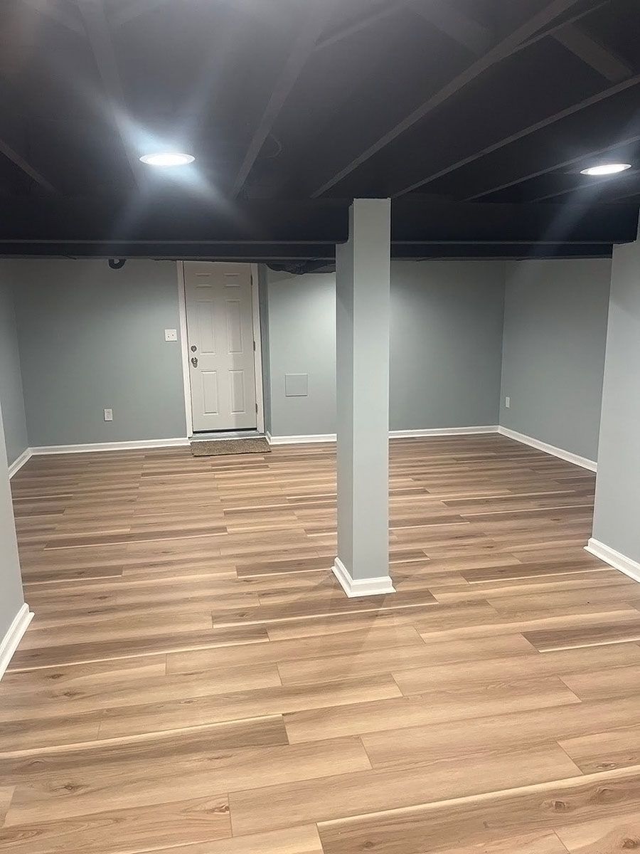 Empty basement room with gray walls, wood-look flooring, exposed ceiling beams, and a central support column
