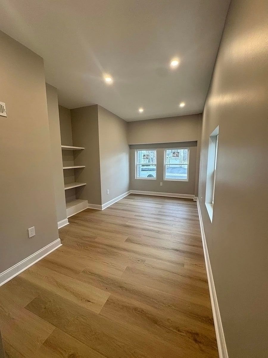 Empty narrow room with wood floors, recessed lights, built-in shelves, and a window at the far end.