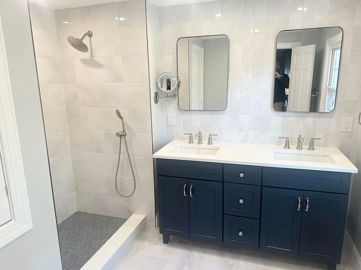 Bright bathroom with walk-in shower and navy double vanity, white tile, and two mirrors.