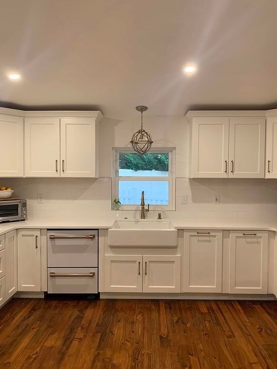 Bright white kitchen with wood floors, cabinets, sink under window, and recessed ceiling lights.