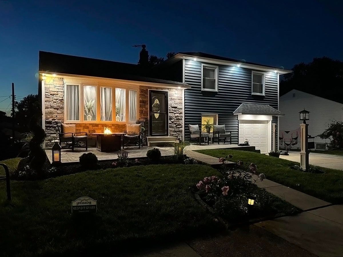 Two-story house at dusk with warm lit windows, stone facade, and a lit front lawn