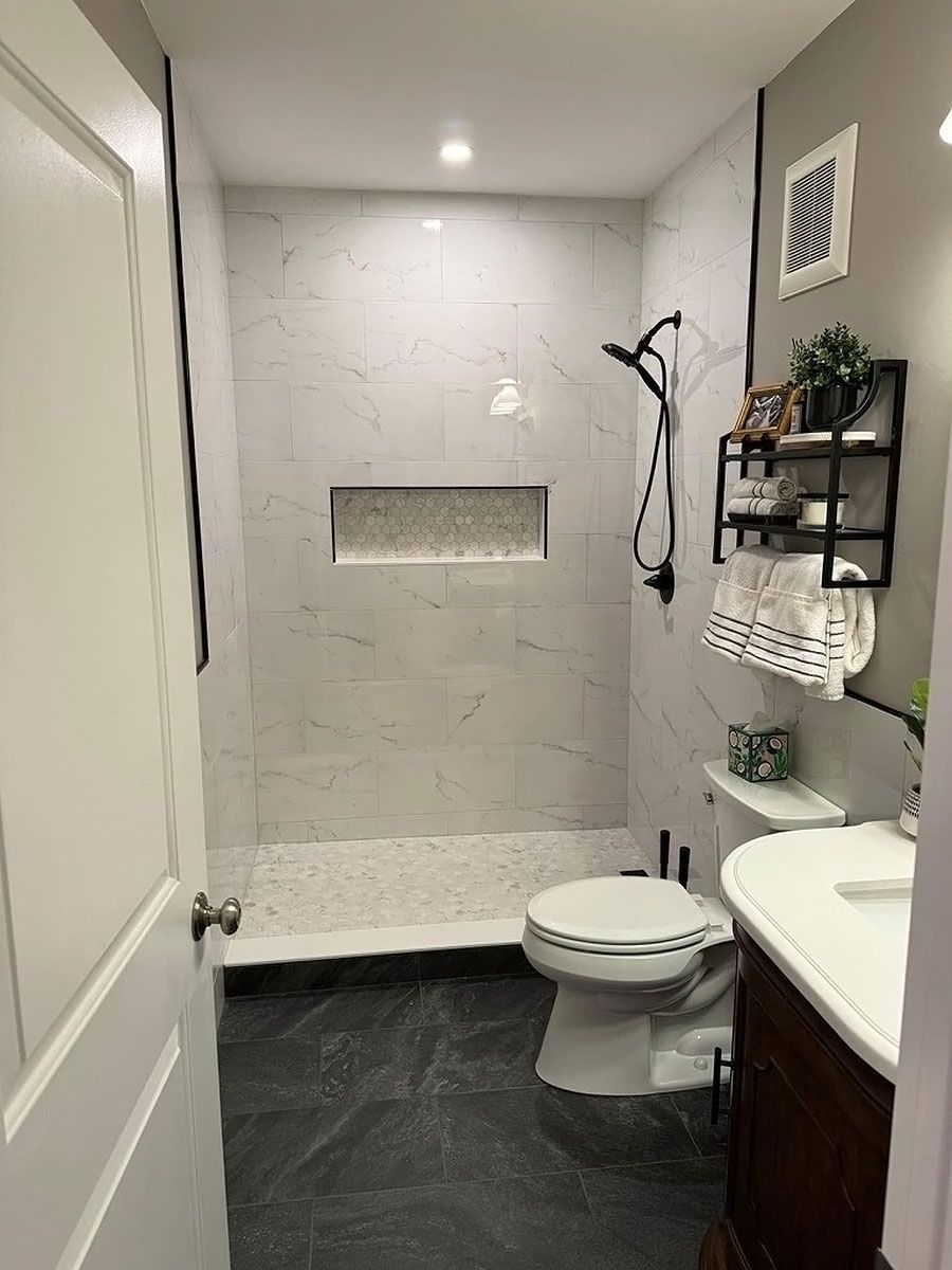 Small white bathroom with walk-in shower, toilet, sink, dark floor, and wall shelves.