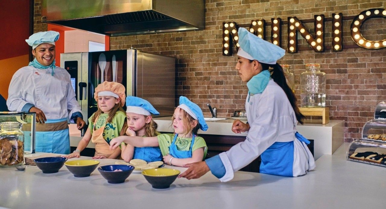 A group of children are cooking in a kitchen with chefs.