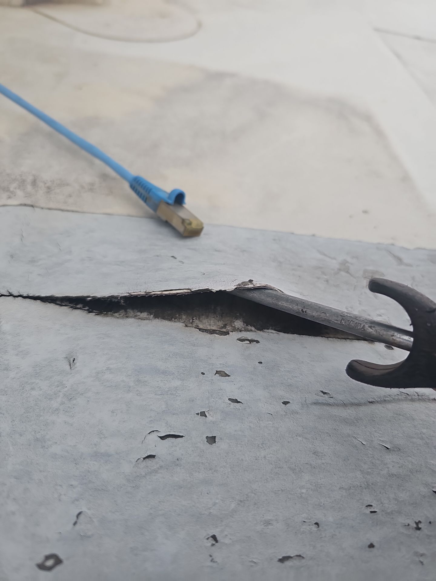 A close-up view showing a metal tool prying open a seam in a grey, textured rooftop surface near a blue wire.