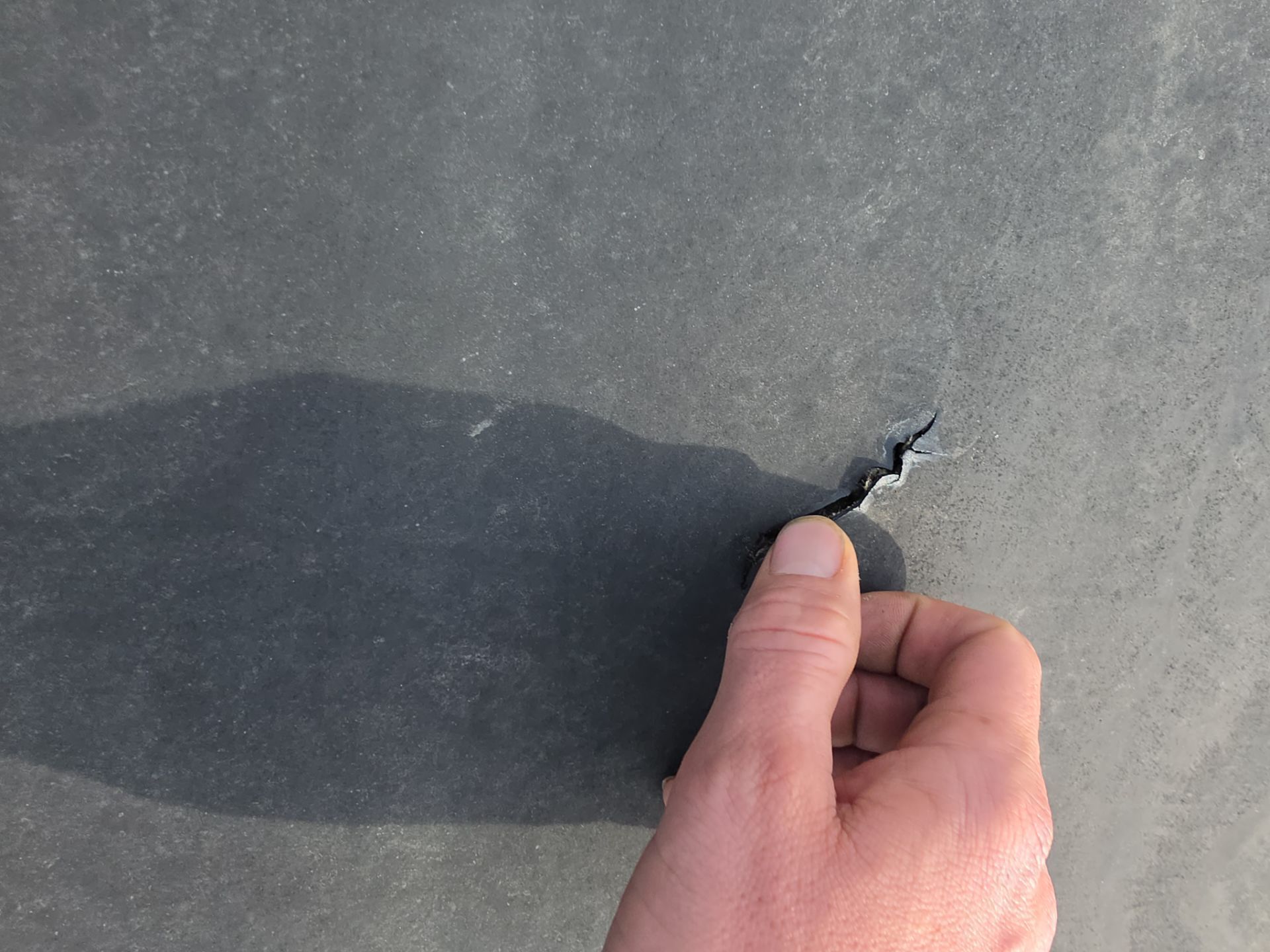 A close-up of a hand lifting the torn, rubbery material of a flat, gray roofing membrane.