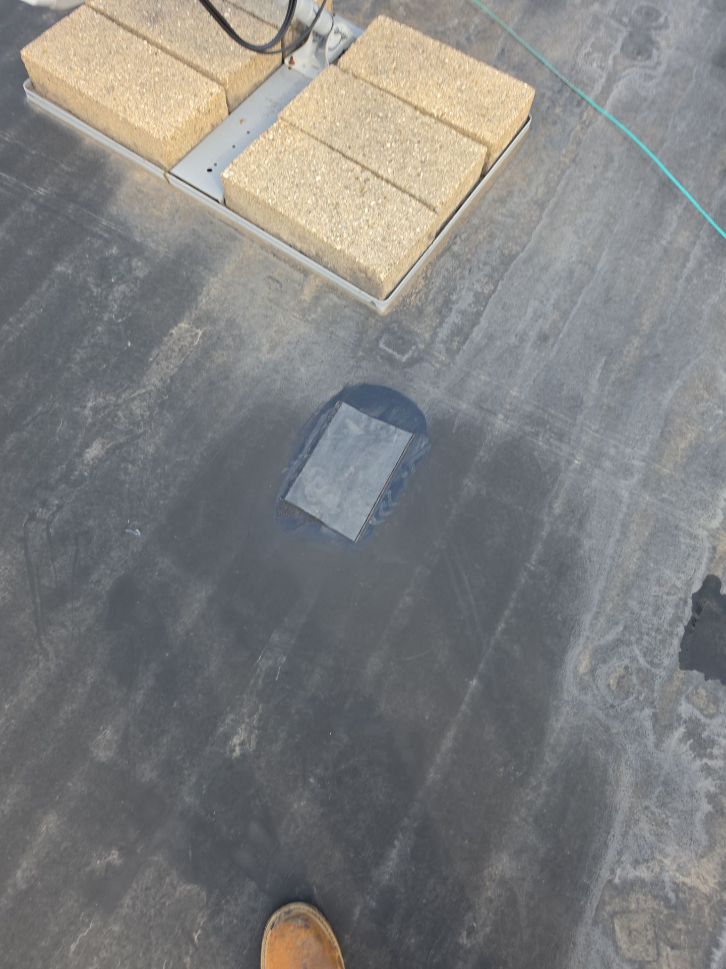 A flat, dark roof surface featuring a metal base weighted by four light-colored bricks, with a small patch repair nearby.