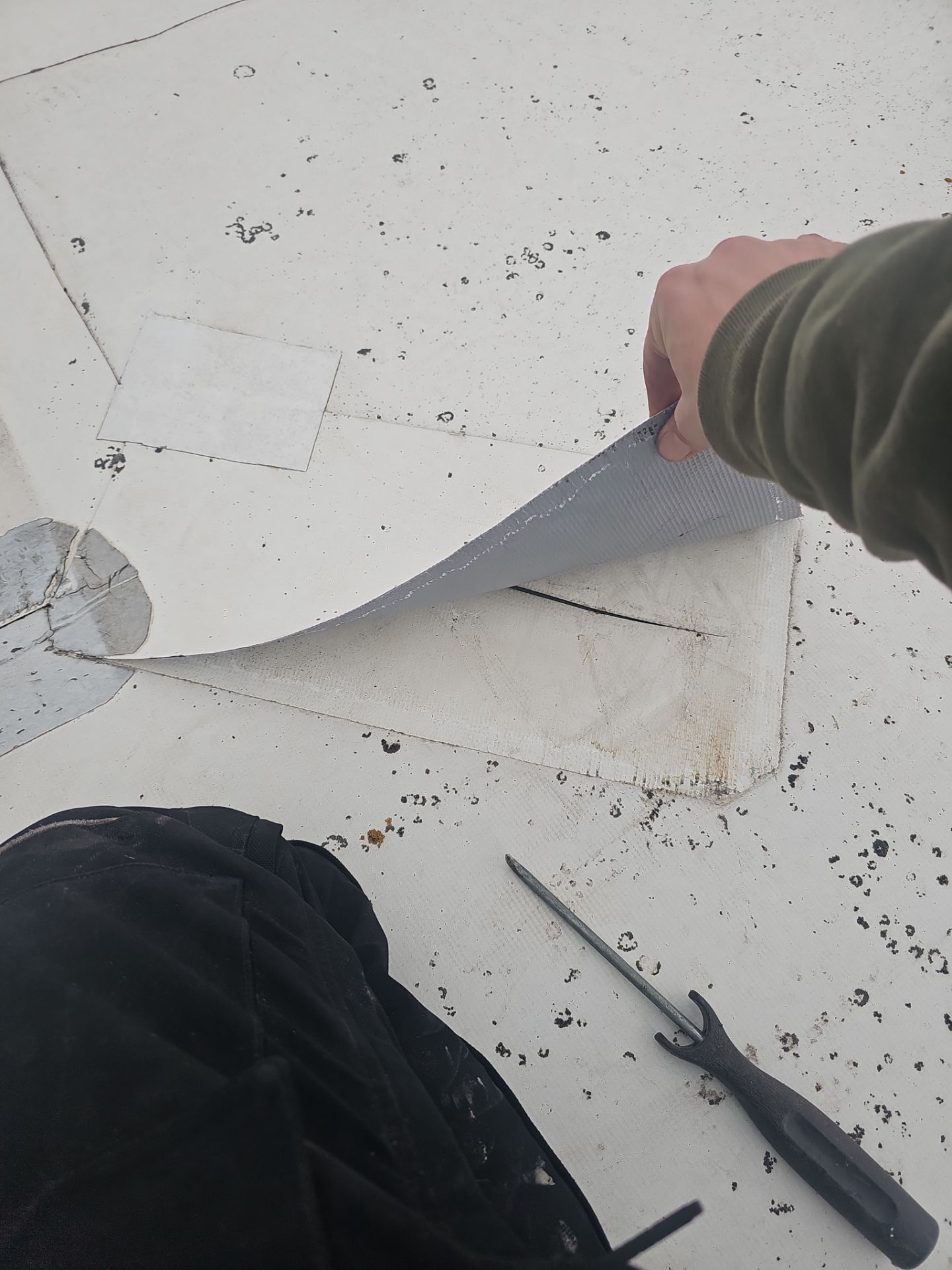 A hand peels back a section of white roofing membrane to reveal a rectangular patch, with a screwdriver resting nearby.