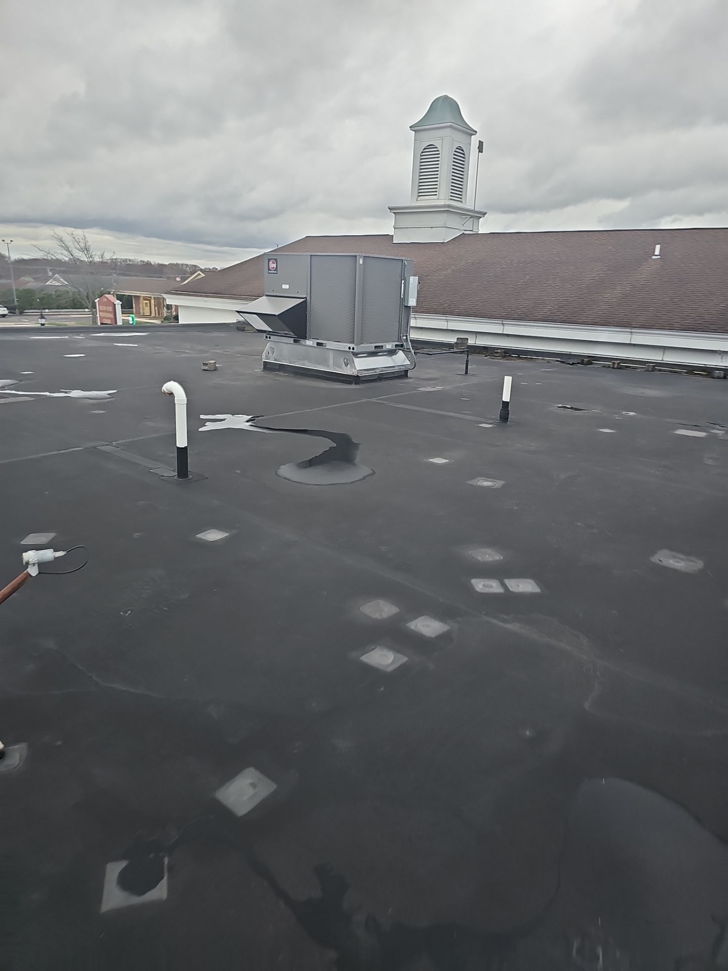 A view of a flat, dark roof featuring a large HVAC unit, several vent pipes, and a building with a cupola in the distance.