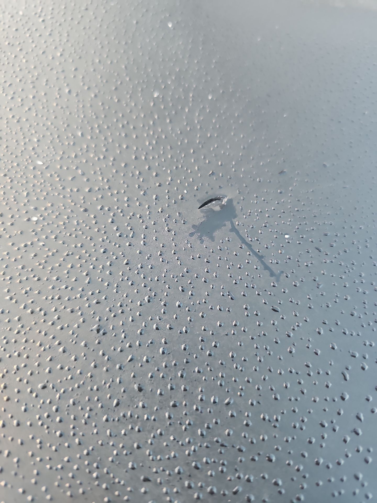 A close-up view of a grey metal surface covered in small water droplets, featuring a small dark scratch in the center.