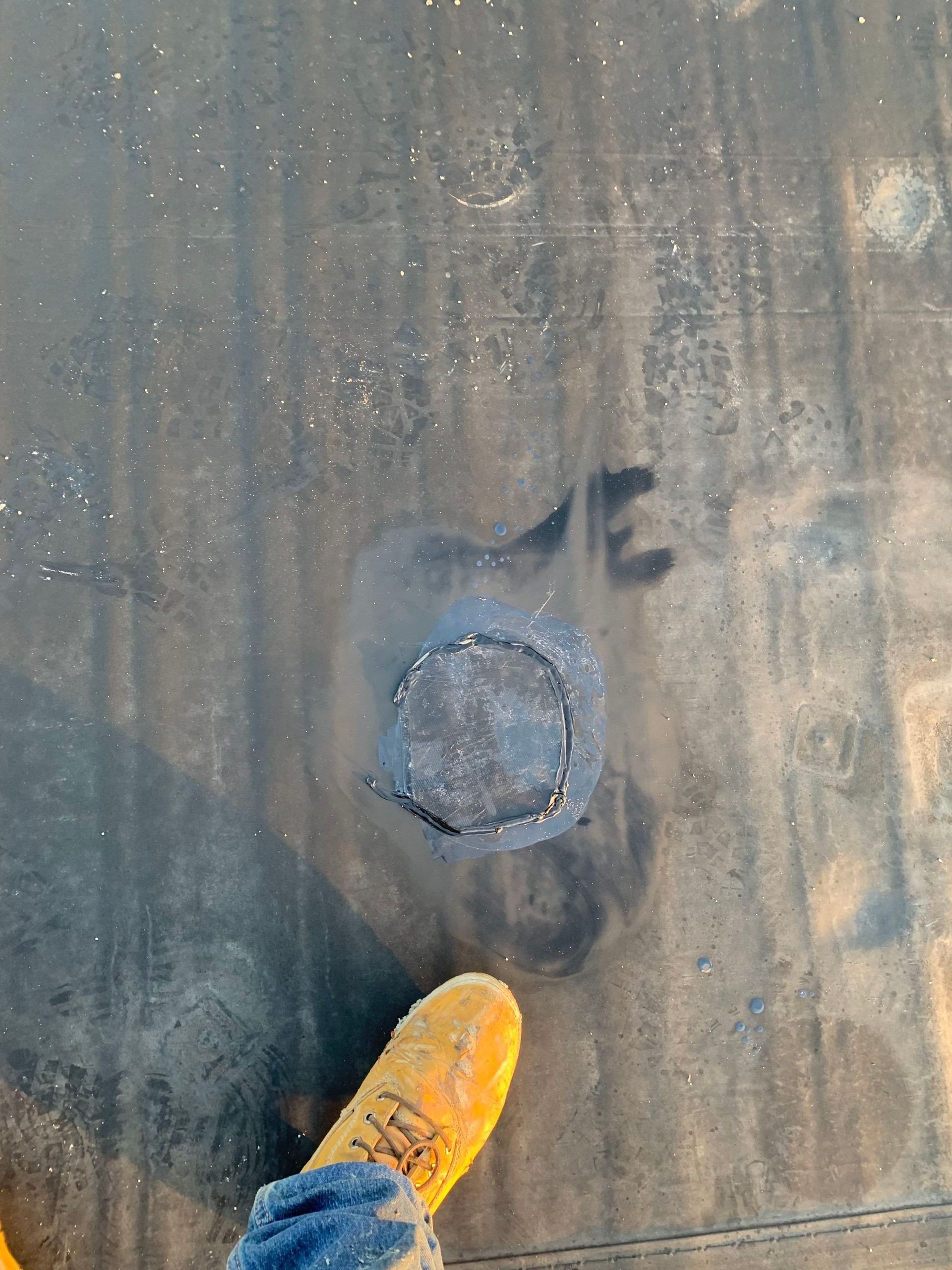 A yellow shoe stands on a dark, flat rooftop next to a circular patch of roofing material marked with black spray paint.