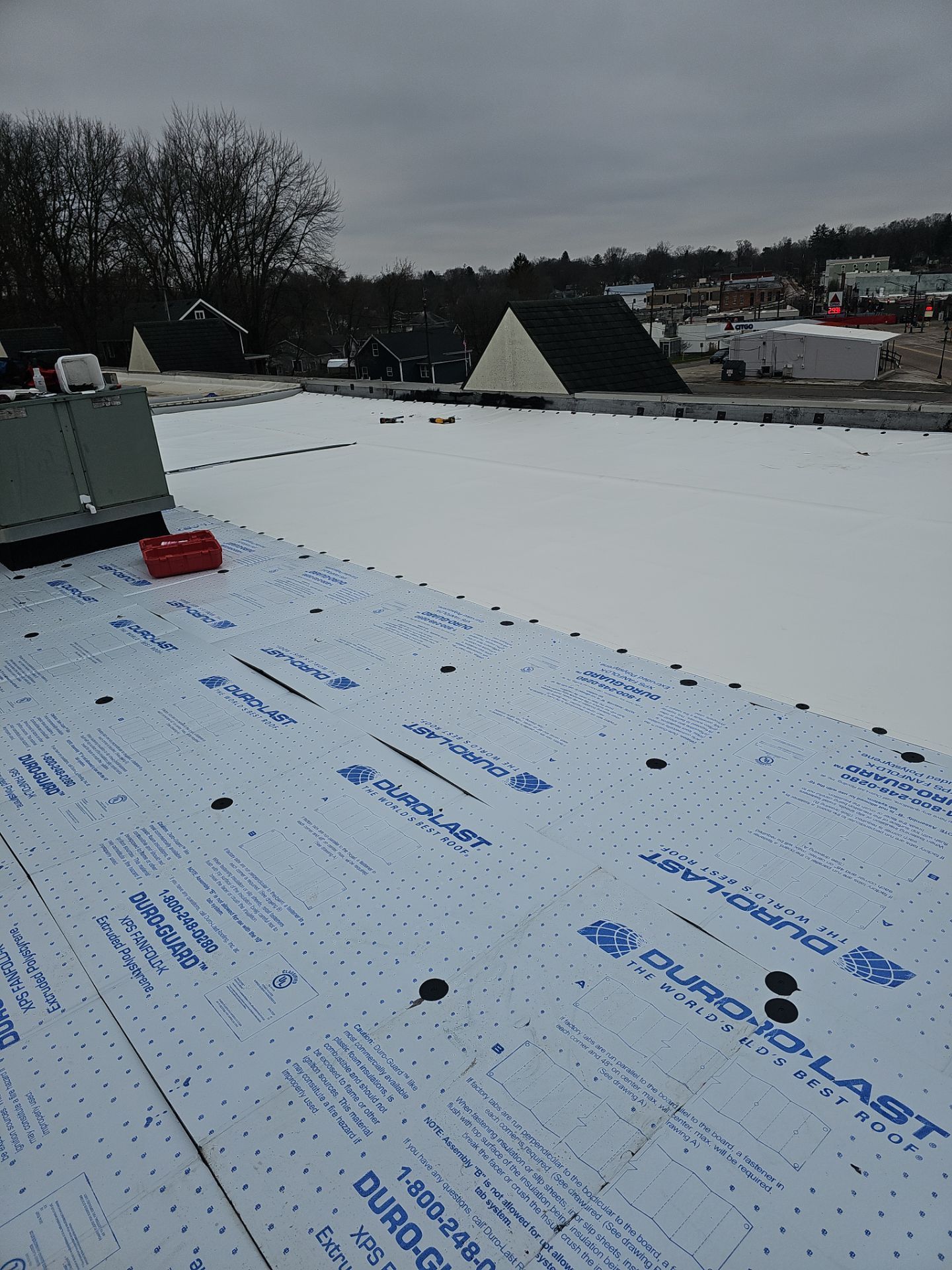 A flat roof under construction, featuring white Duro-Last insulating boards with fasteners, under a cloudy sky.