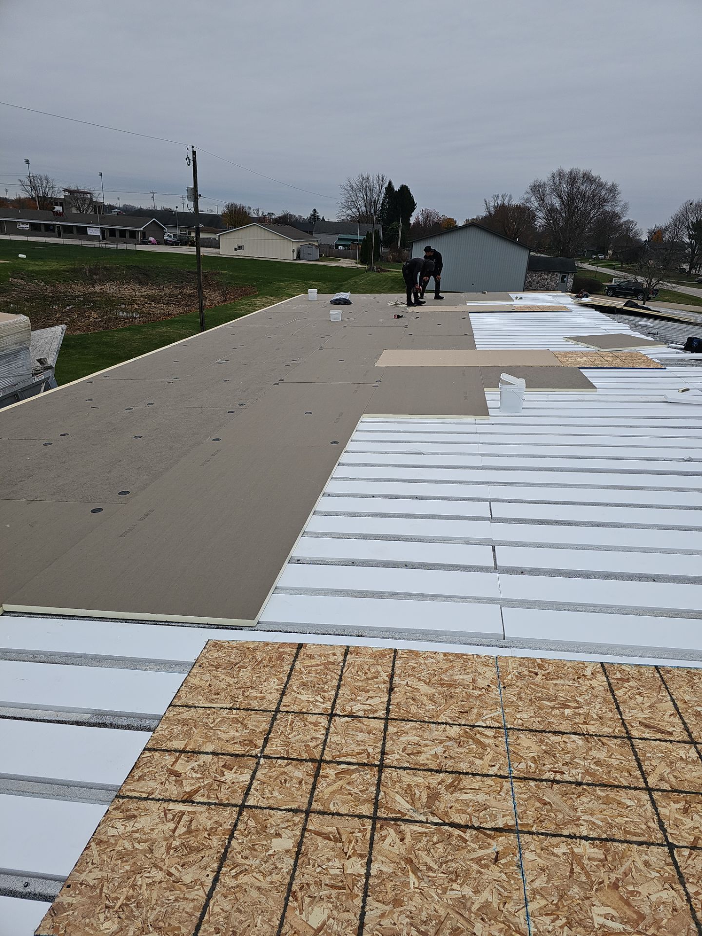 Construction workers install plywood panels on a flat roof under an overcast sky.