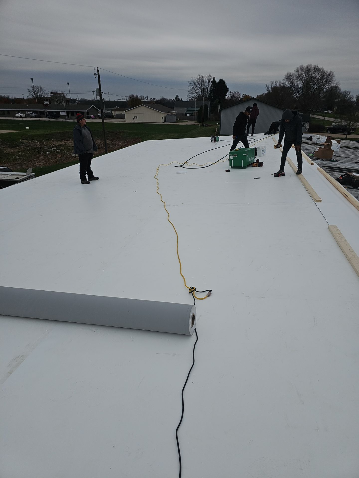 Workers install a white TPO roofing membrane on a flat roof under an overcast sky.
