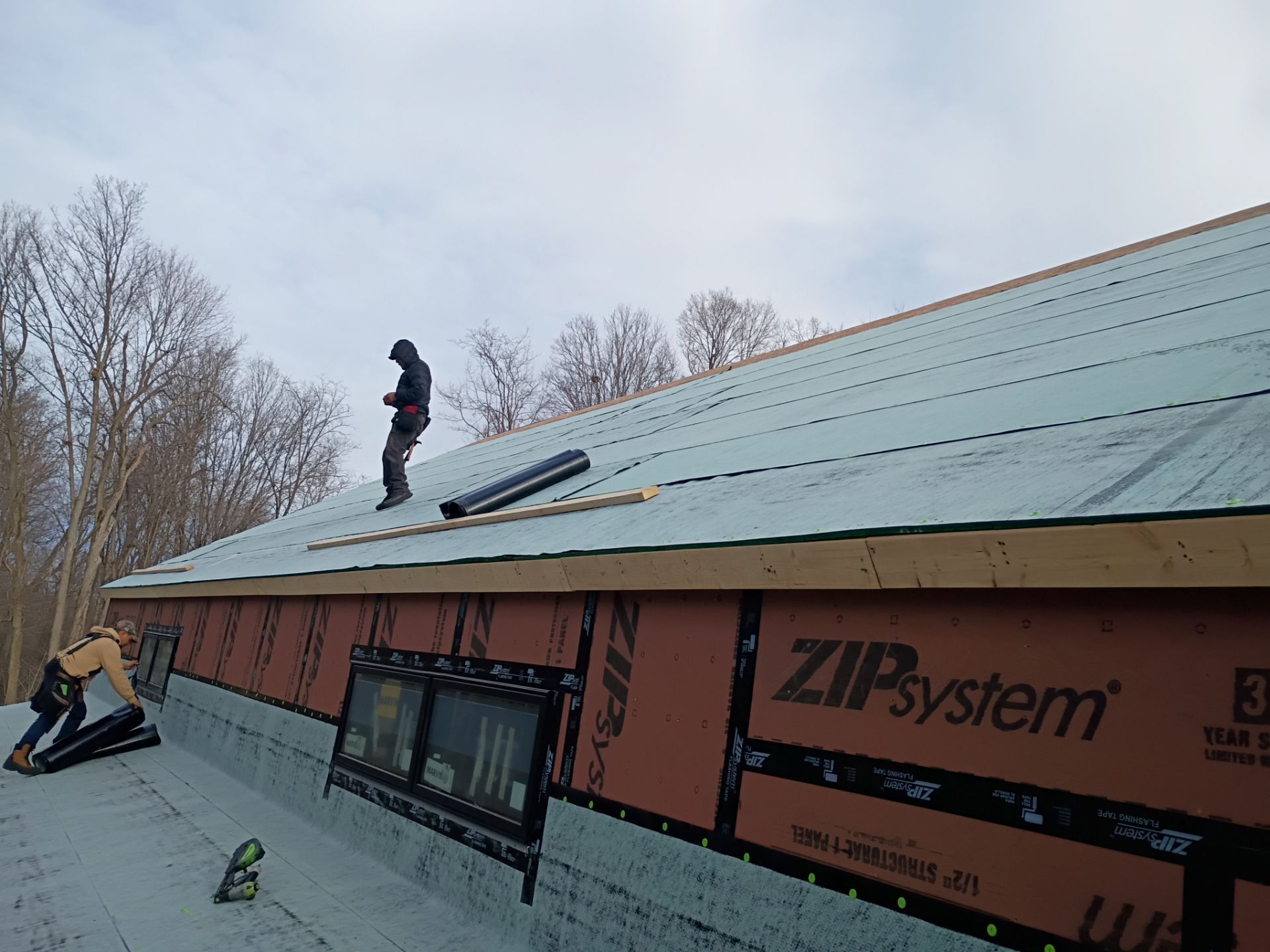 Two workers in workwear install roofing materials and ZIP system wall panels on a house under construction.