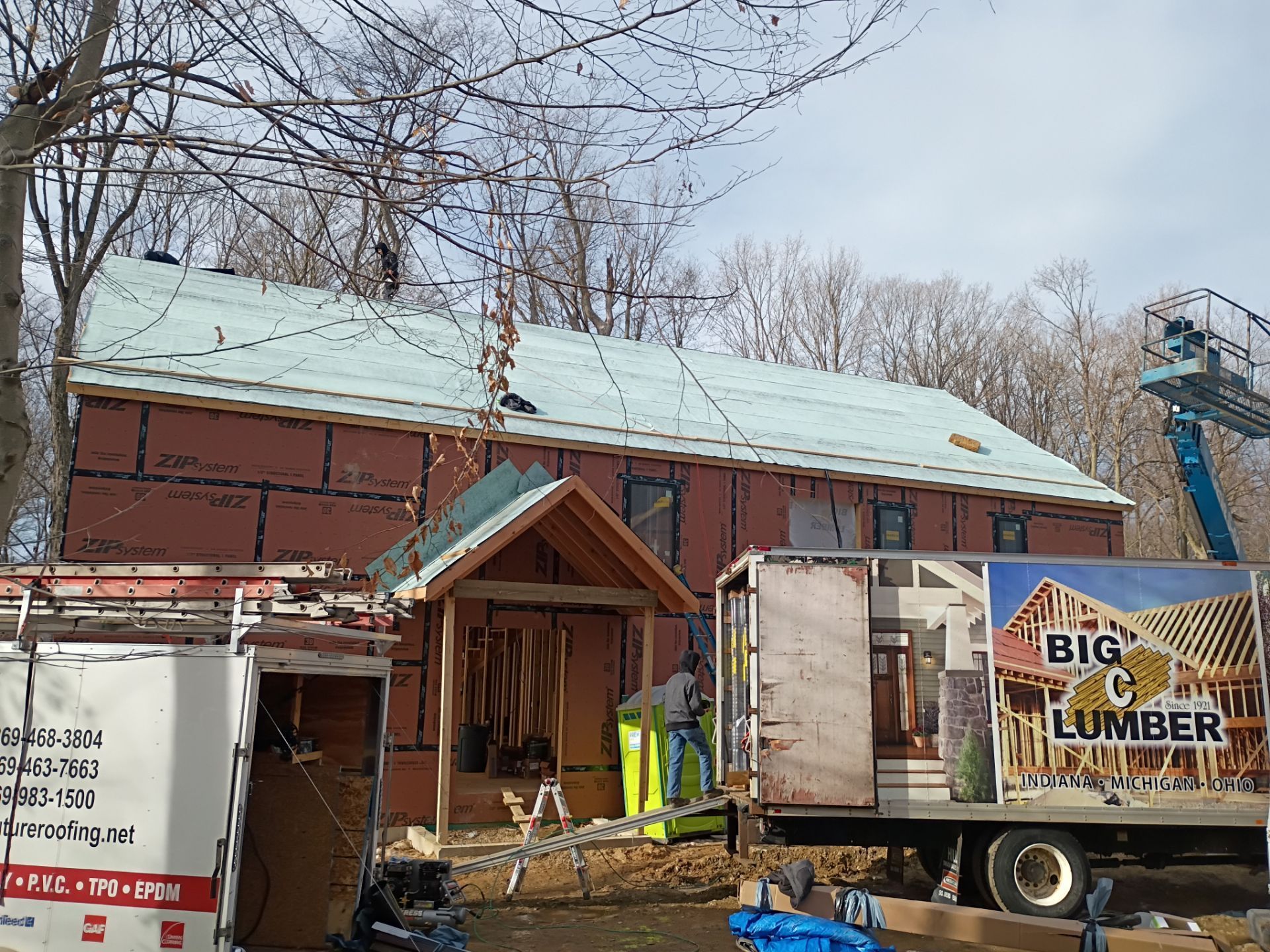 A house under construction with red siding and a partially shingled roof, flanked by work trucks and a boom lift.
