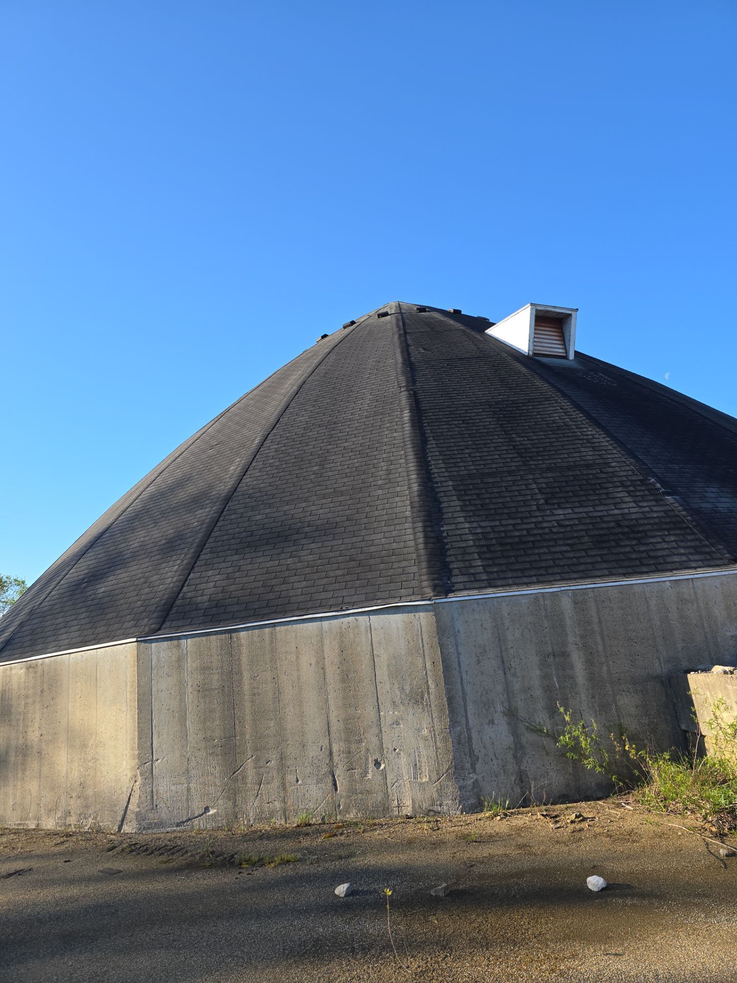 A large, conical storage dome with a dark, shingled roof and concrete base under a clear blue sky.