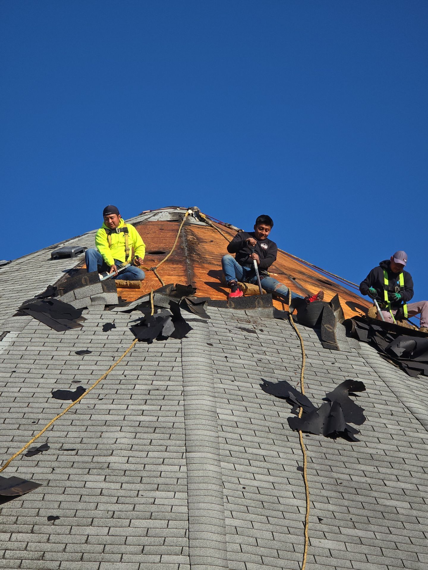 Three roofers in safety gear work on a steep, shingled roof under a clear blue sky.