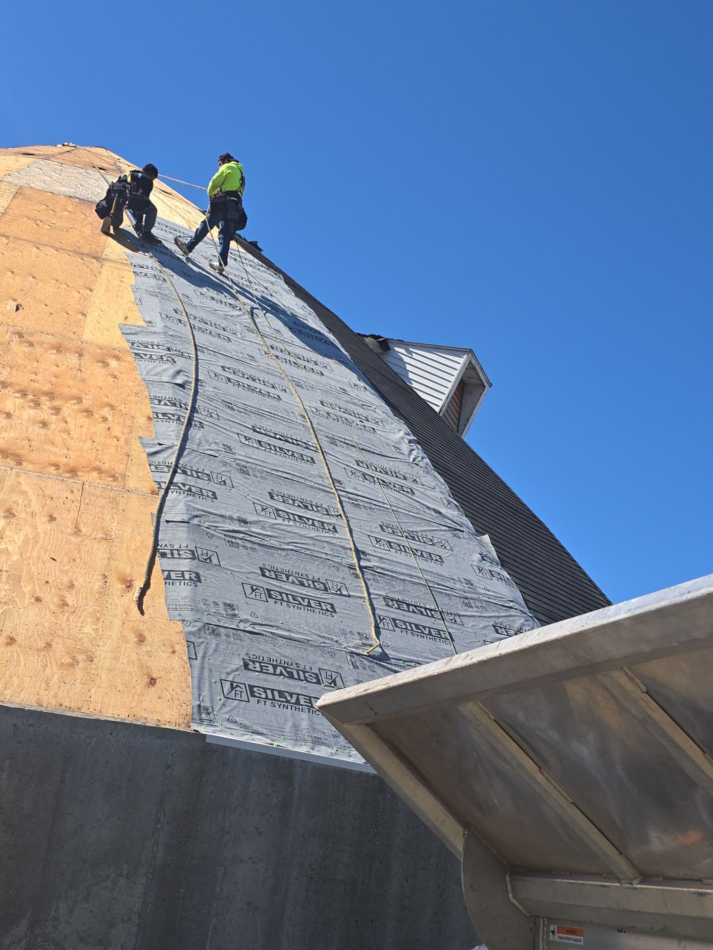Two workers on a steep, pitched roof installing grey underlayment over exposed wooden roof decking under a clear blue sky.