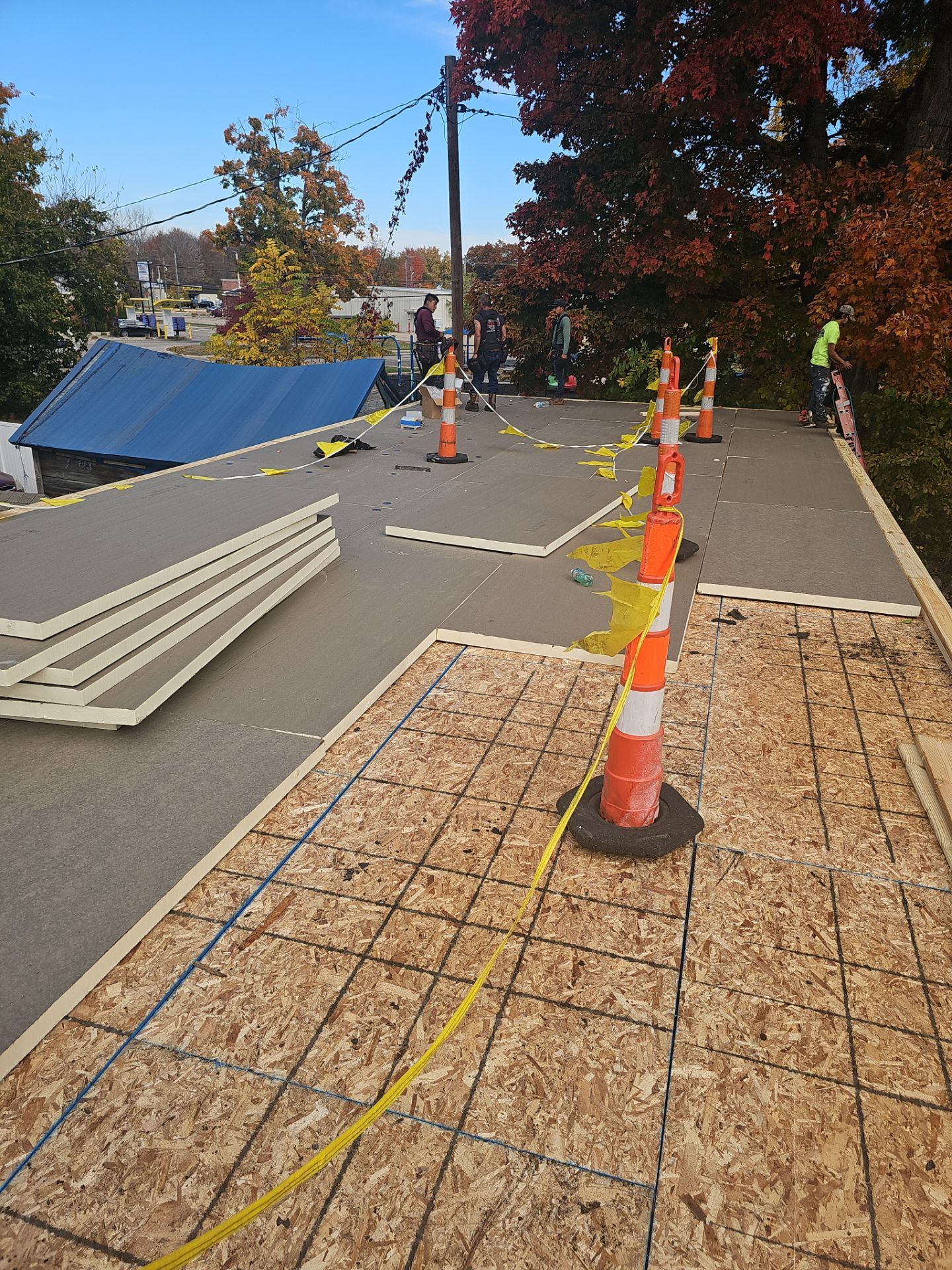 Construction workers installing plywood roof decking on a sunny day, marked by orange safety cones and yellow caution tape.
