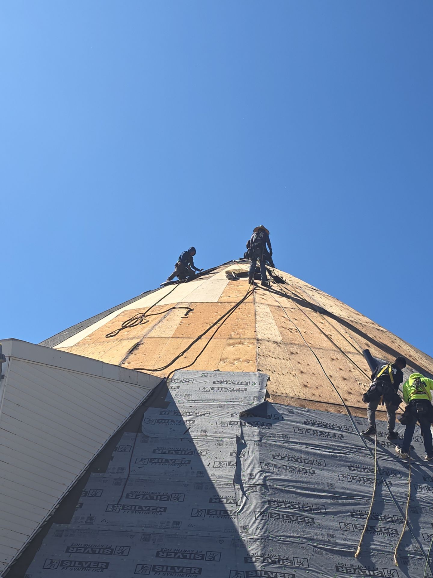 Roofers work on a steep, partially shingled roof under a clear blue sky.