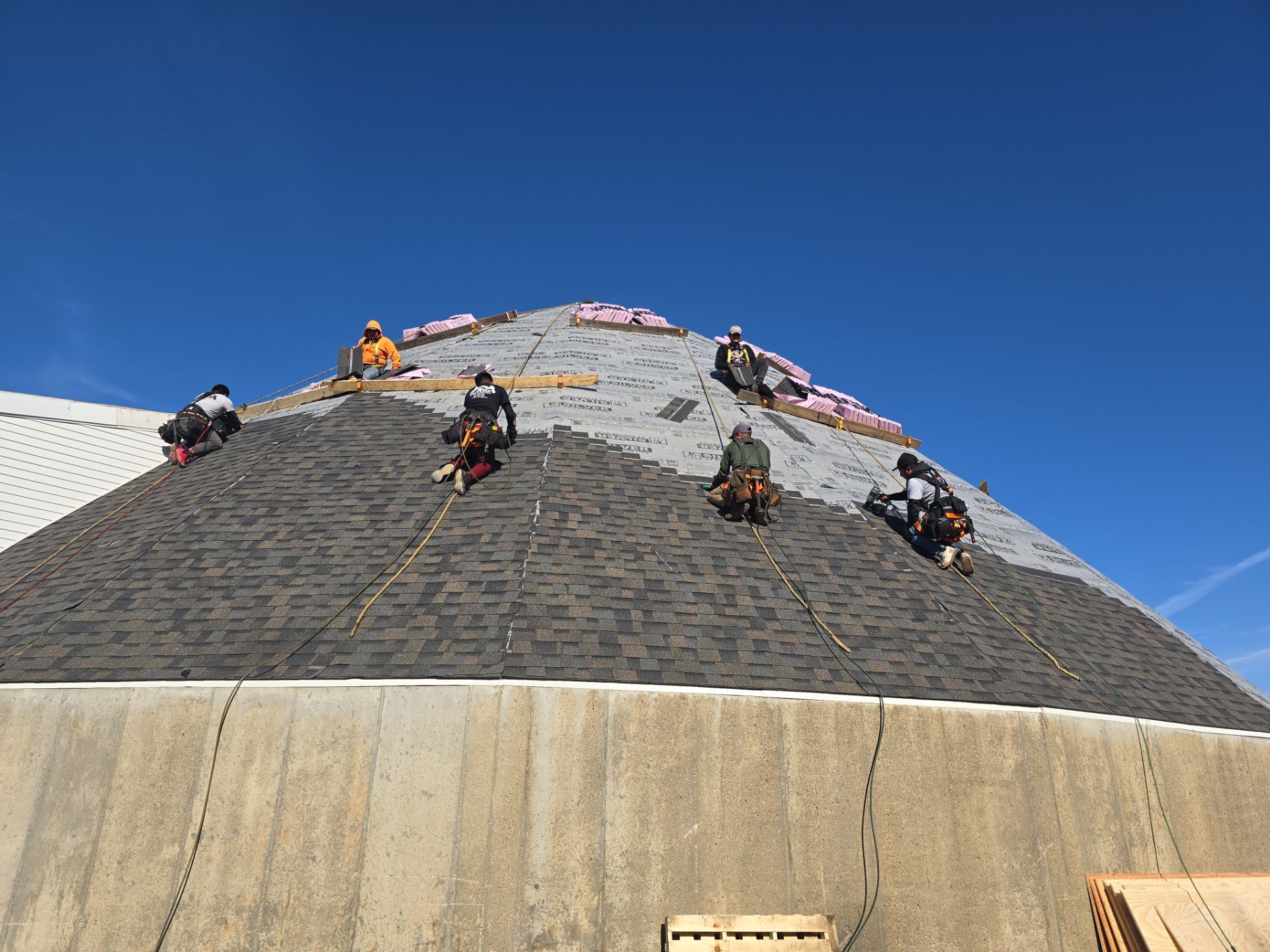 Workers install asphalt shingles on a steep, conical roof under a clear blue sky.