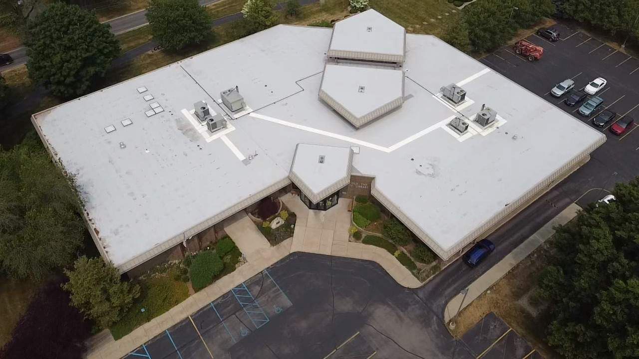 Aerial view of a flat-roofed commercial building with a grey roof, surrounding trees, and a parking lot with several cars.