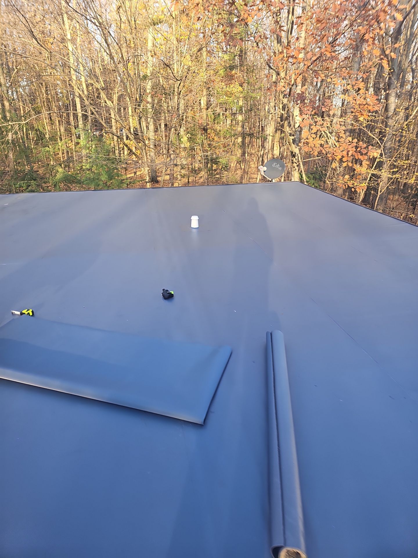A smooth, gray roof surface with a small white pipe and a rolled-up edge, set against a background of autumn trees.