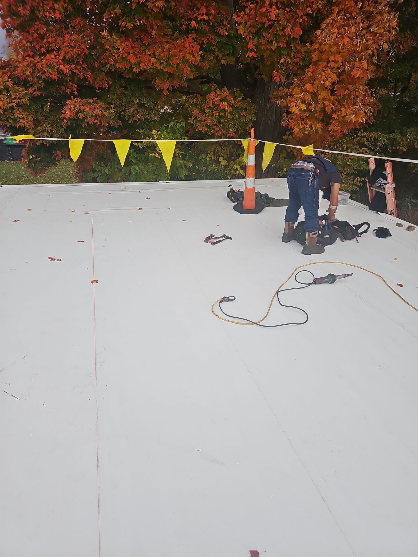 A worker installs a white roof membrane on a building site marked by orange safety cones and yellow caution tape.