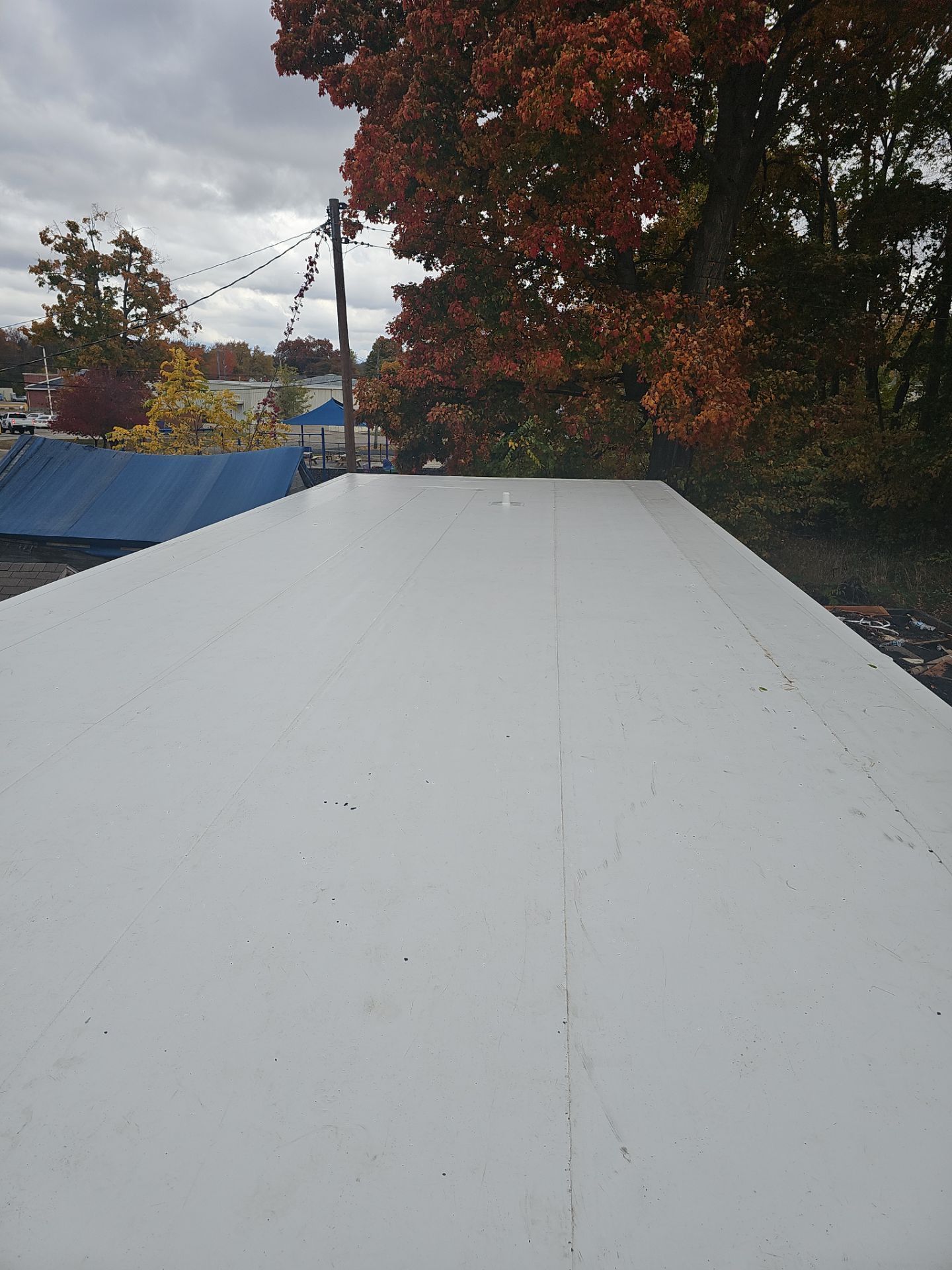 An elevated view of a flat white rooftop against a backdrop of trees with autumn foliage under a cloudy sky.