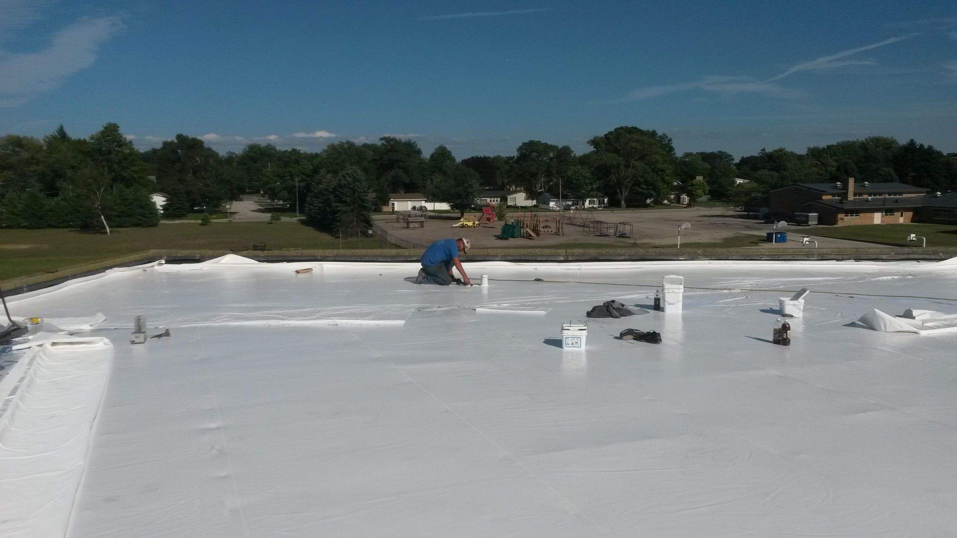 A worker applies white roofing material to a flat roof on a sunny day.