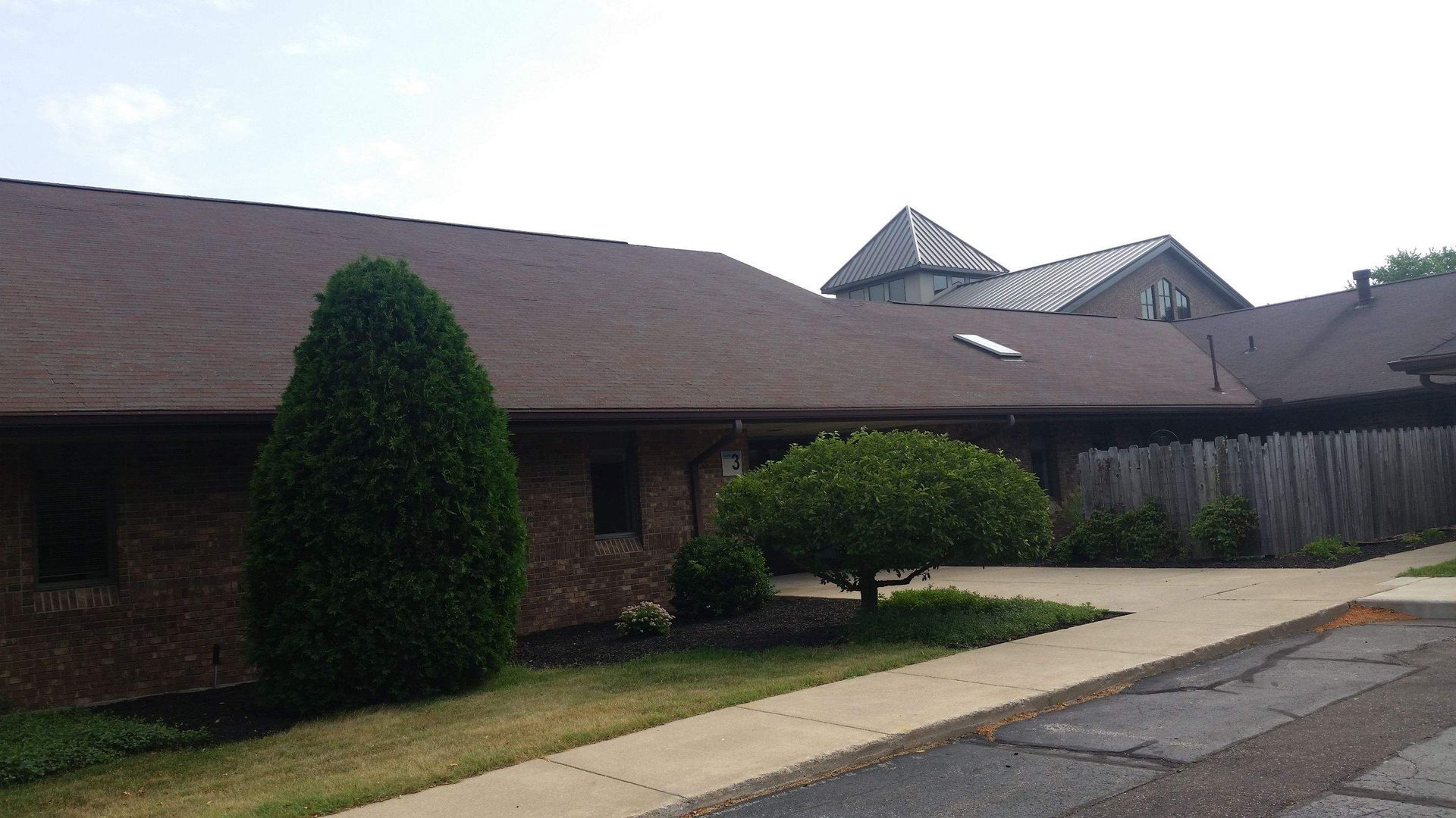 A low-profile brick building with a brown shingled roof, a paved walkway, and manicured landscaping under a cloudy sky.