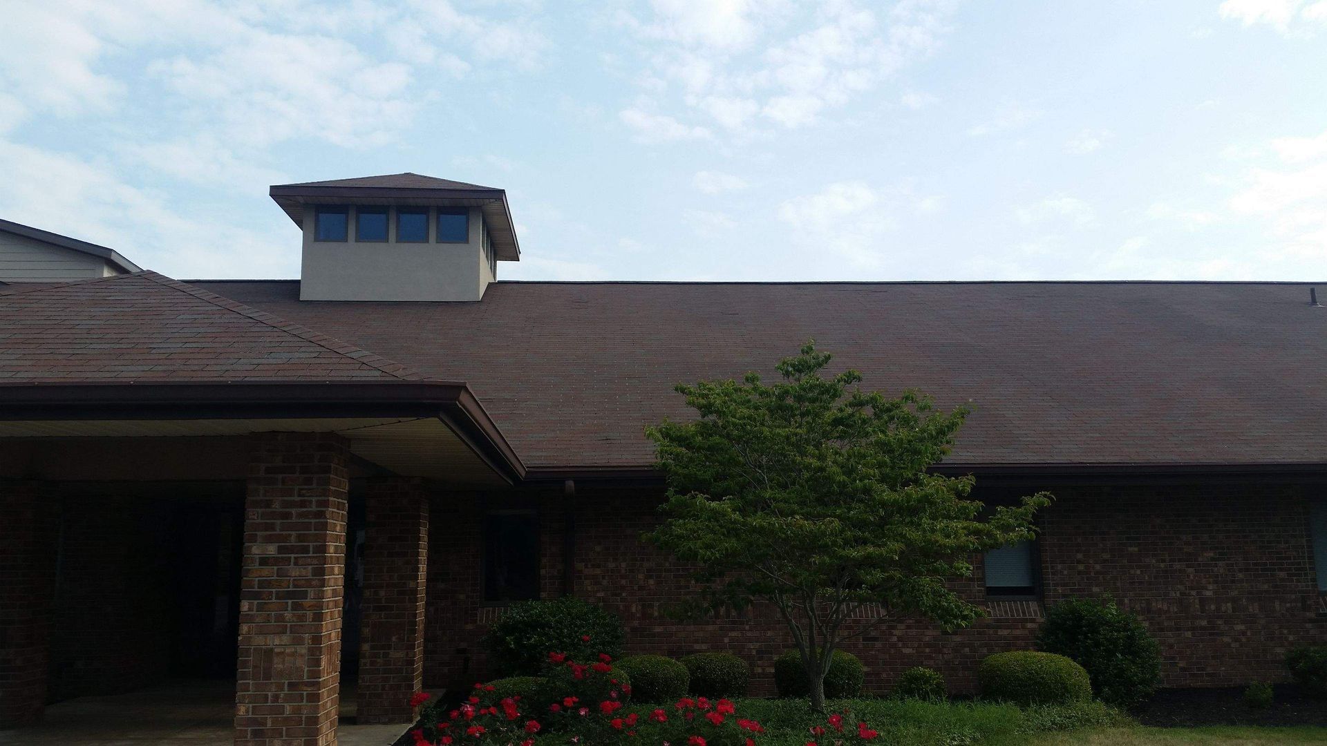 A brick building with a dark shingled roof and a central cupola sits behind a small green tree and red flowers.