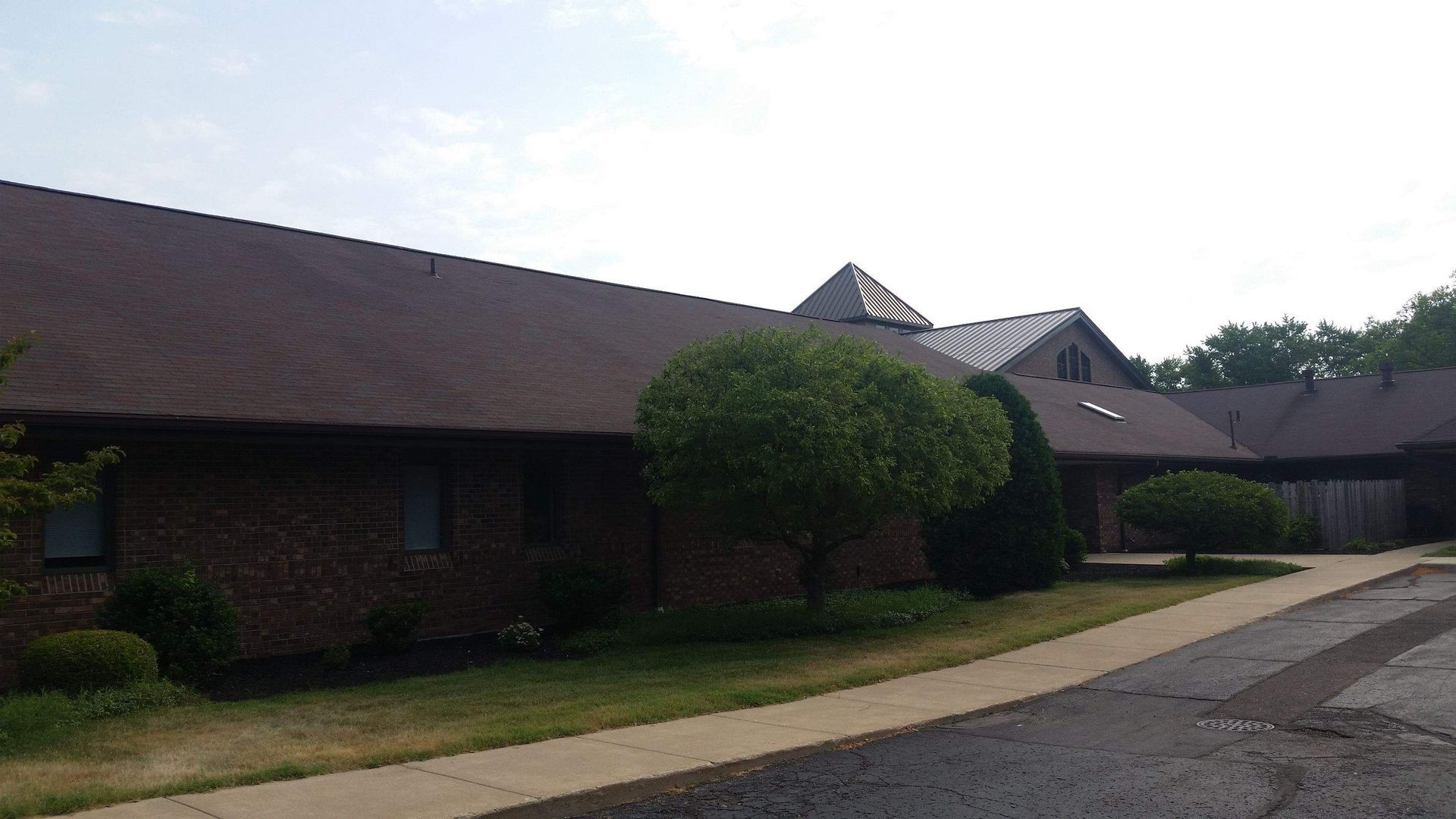 A brick building with a dark shingled roof, a central pyramid skylight, and a paved walkway in front.