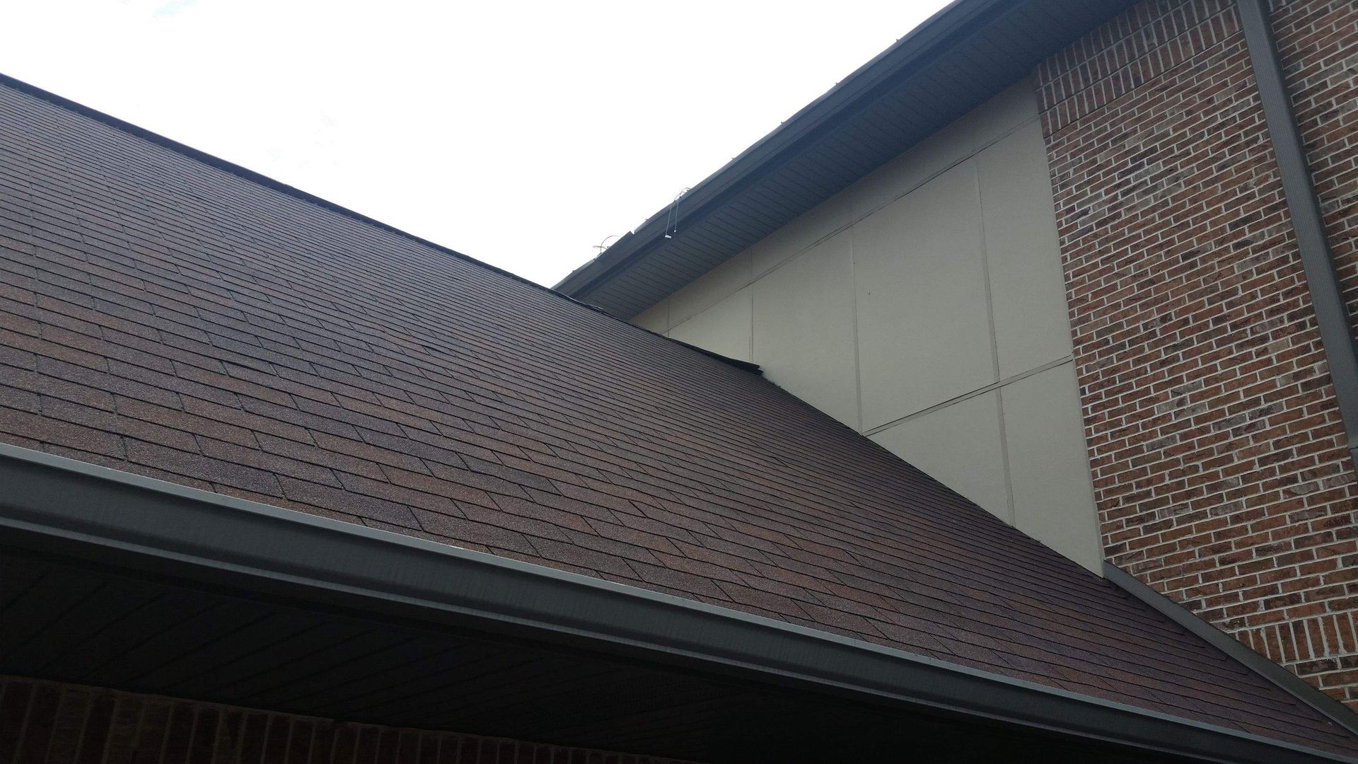 A sloped roof with dark asphalt shingles meets a light-colored exterior wall and a vertical brick column.