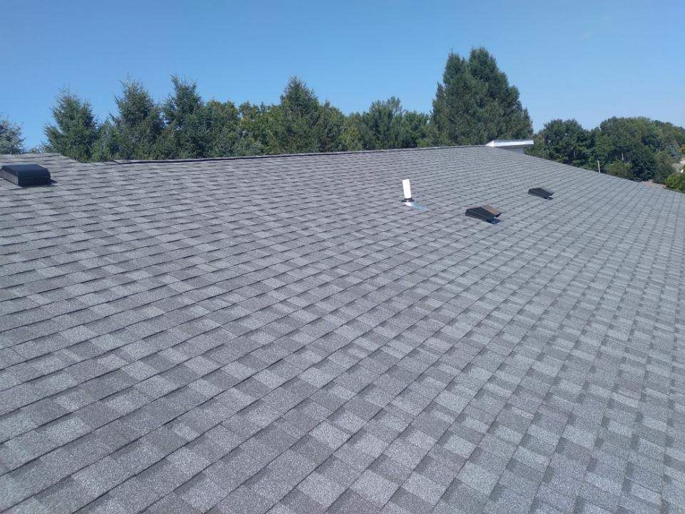 A grey asphalt shingle roof under a clear blue sky, featuring a ventilation pipe and several dark roof vents.