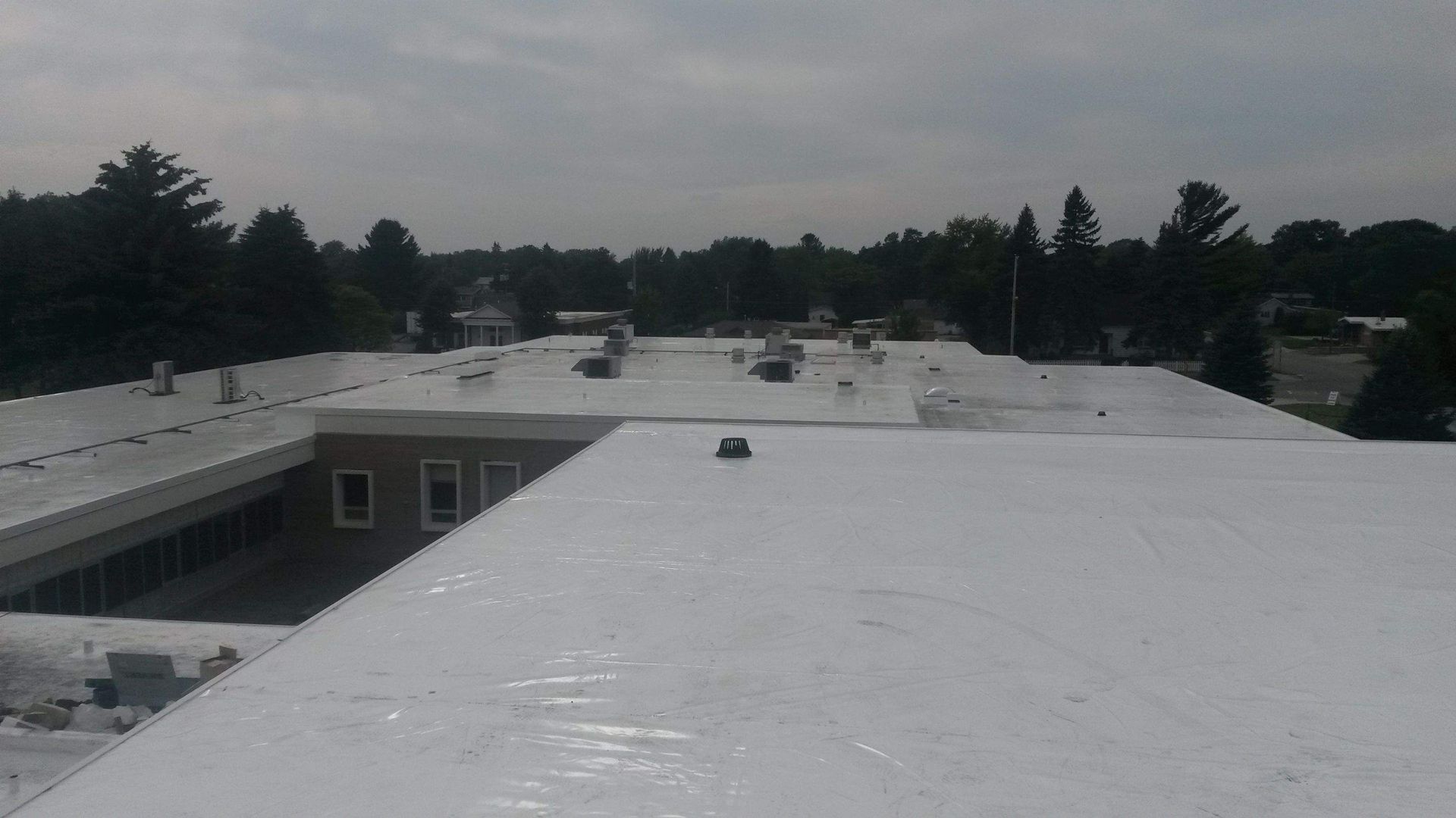 A wide-angle, elevated view of a flat, white-coated commercial building roof surrounded by green trees under a gray sky.