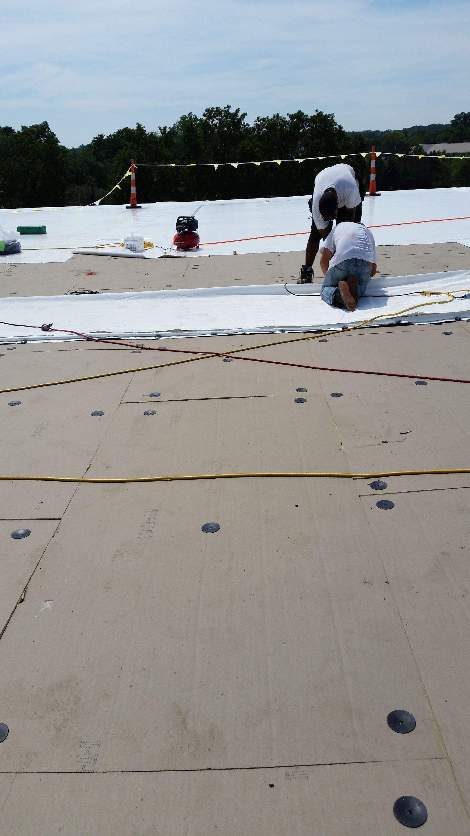 Two workers install a white roofing membrane on a flat, tan rooftop surface.