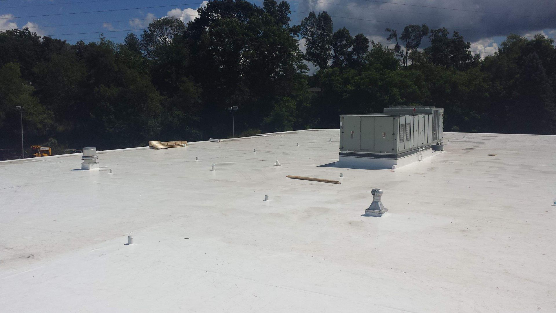 A flat white roof features an HVAC unit and several small vents, set against a backdrop of green trees under a blue sky.