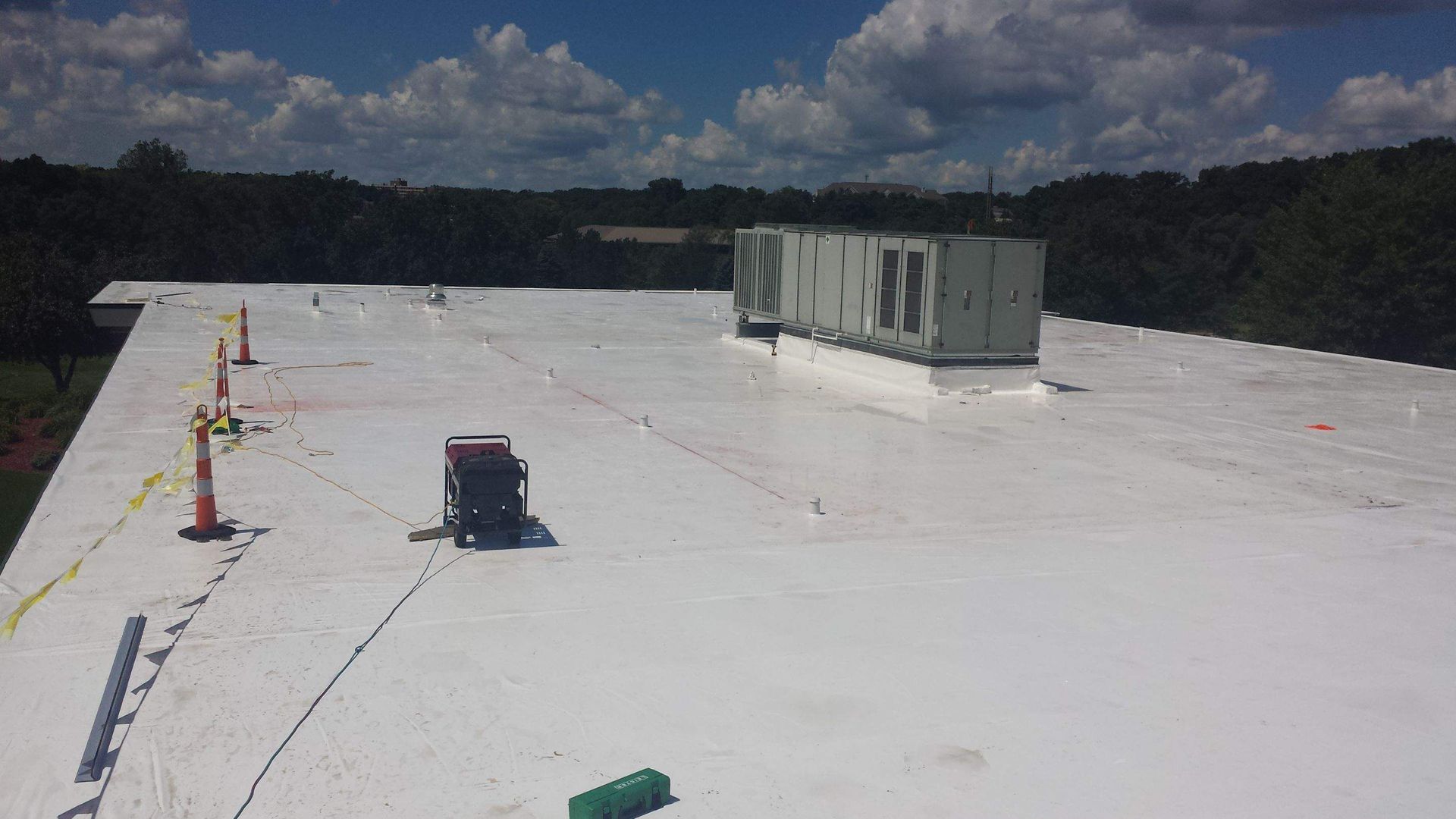 A wide, flat roof with a white coating, a large gray HVAC unit, and several orange safety cones under a blue cloudy sky.